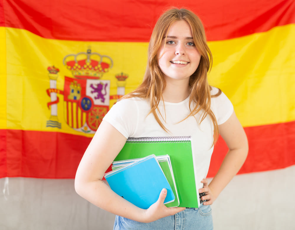 Happy smiling teen girl holding spiral notepads under her arm against of national flag of Spain