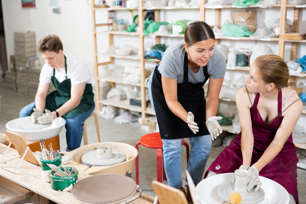 Young woman teacher helps teenagers boy and girl to make product on potter's wheel