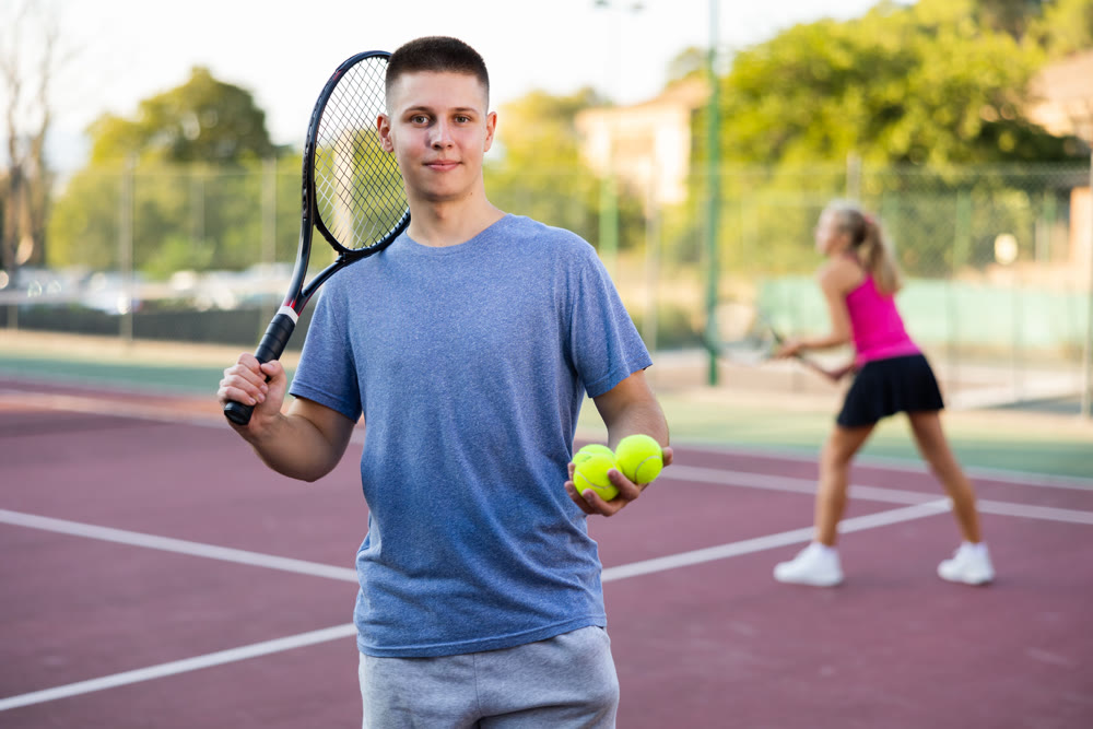 Young man in polo shirt carrying tennis racket on shoulder standing on tennis court