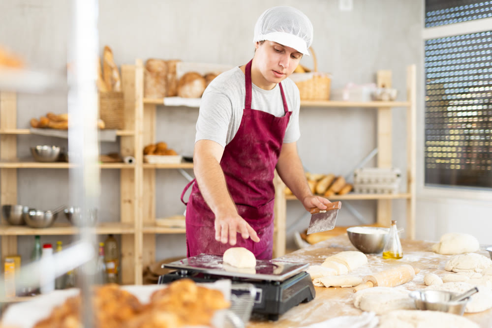 Male cook is working with dough, weighing piece of dough on scale.