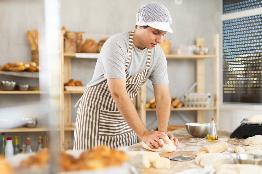 Guy baker in uniform prepares raw dough, kneads it with hands