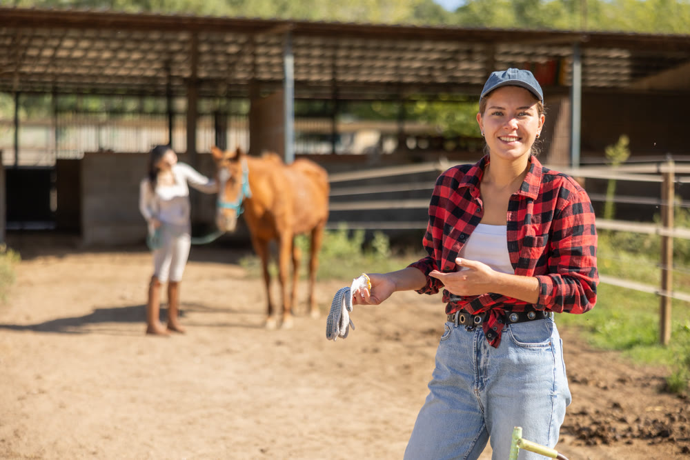 Positive female stable worker stands near wooden fence of paddock and watches horse.