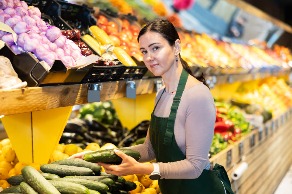 Middle-aged female seller putting cucumbers on counter in grocery market