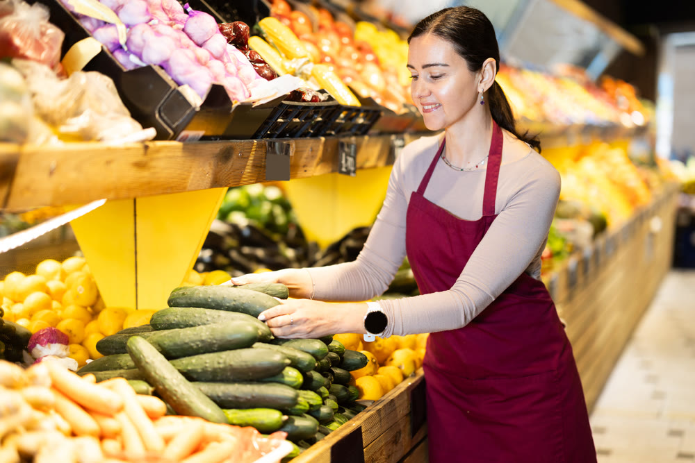 Female salesperson carefully places ripe cucumbers on counter of a grocery supermarket