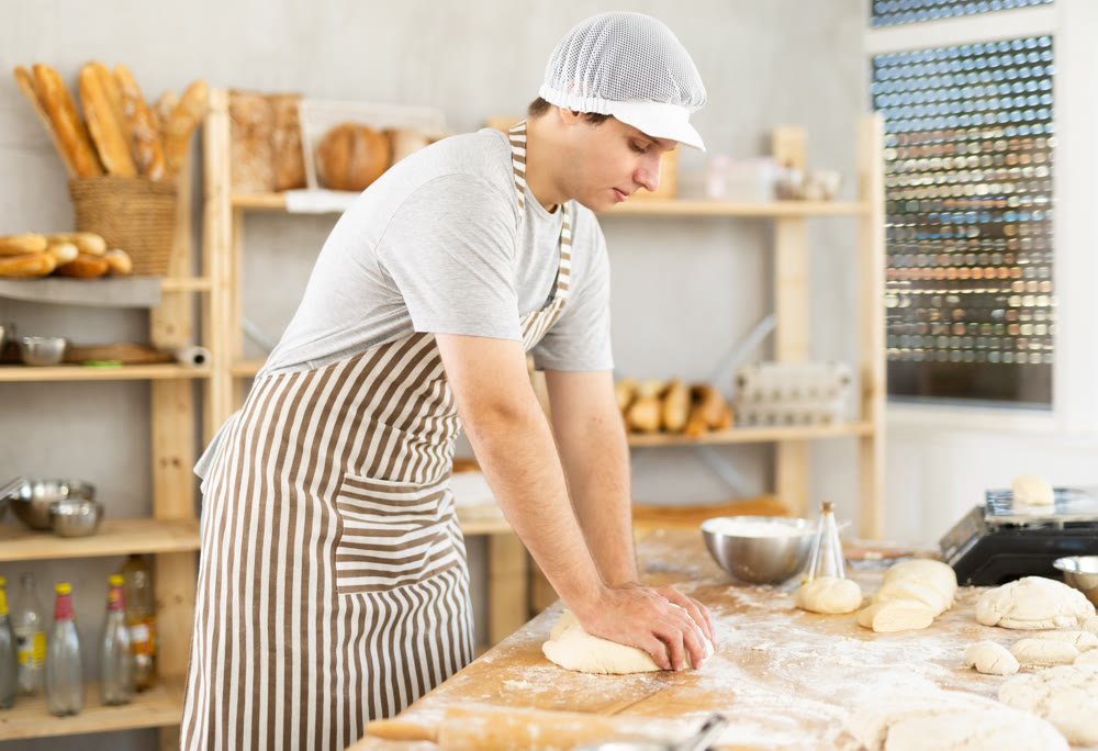 Portrait of young guy baker in apron working with dough, kneading and forming bakery products in kitchen in bakery