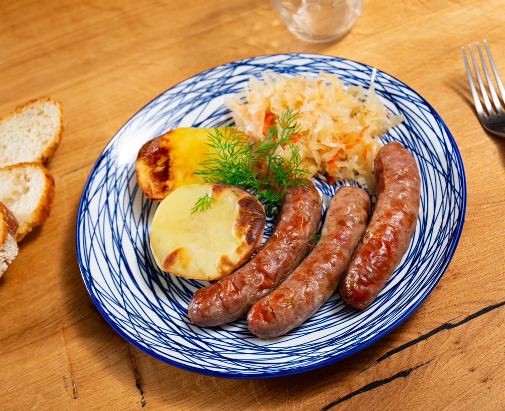 On table there is plate with grilled sausages, sauerkraut and slices of potatoes