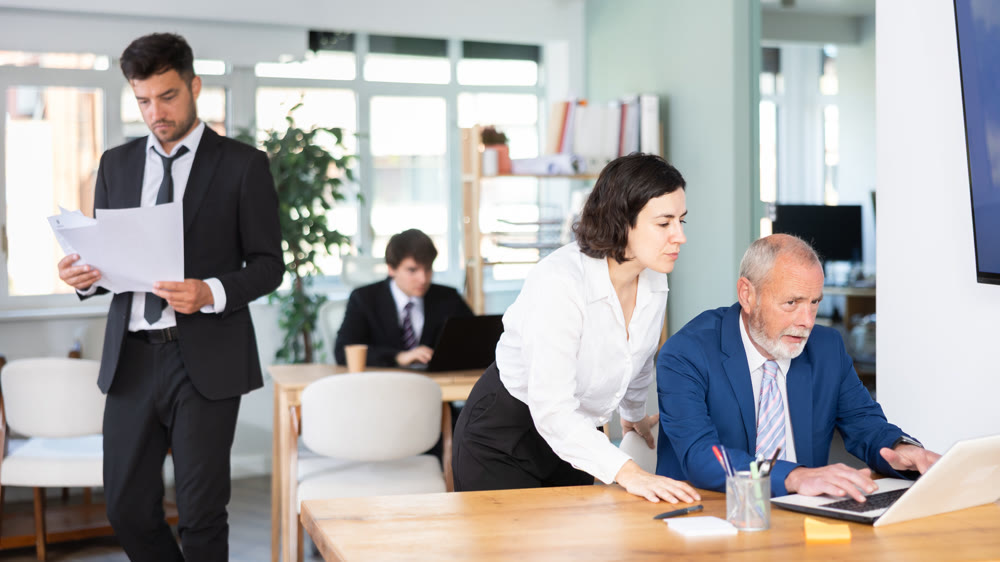 Young female business worker helping mature male colleague with laptop, discussing new project in office