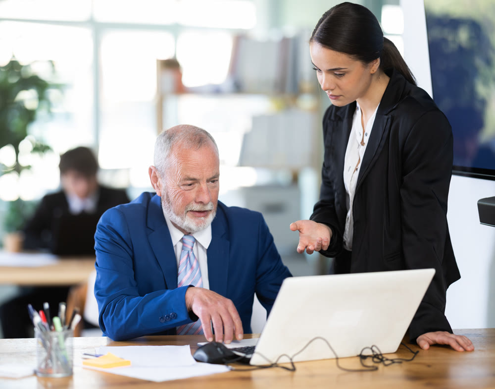 Young female business worker helping mature male colleague with laptop, discussing new project in office