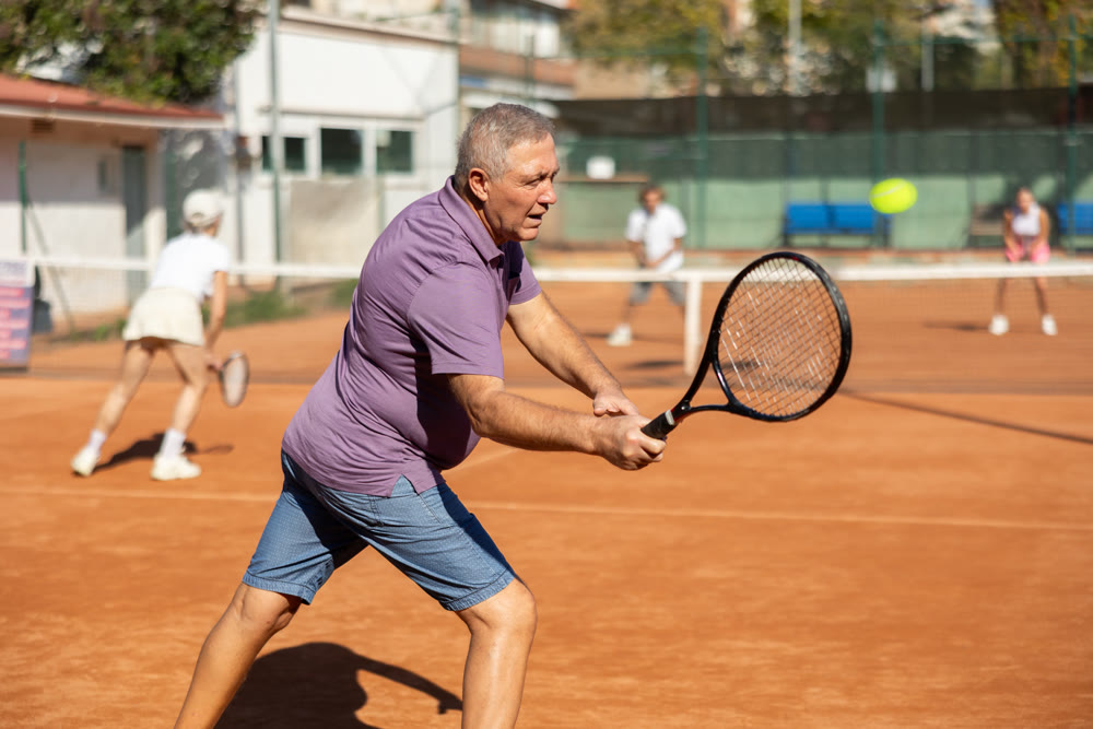 Elderly couple in sportswear spending time playing sports on tennis courts, old man receiving hit from tennis racket