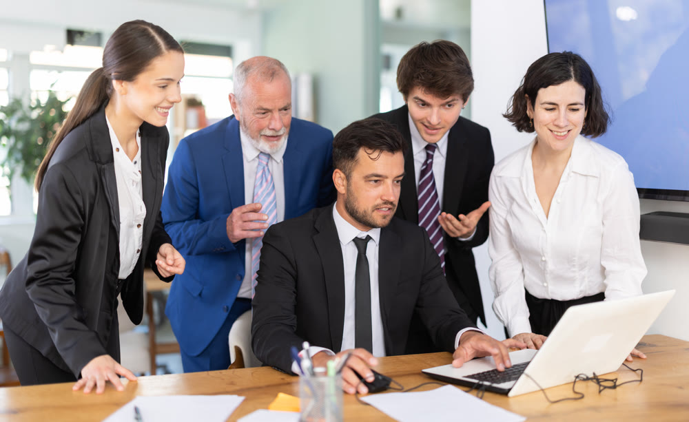Group of colleagues looking at adult man's laptop and laughing in office