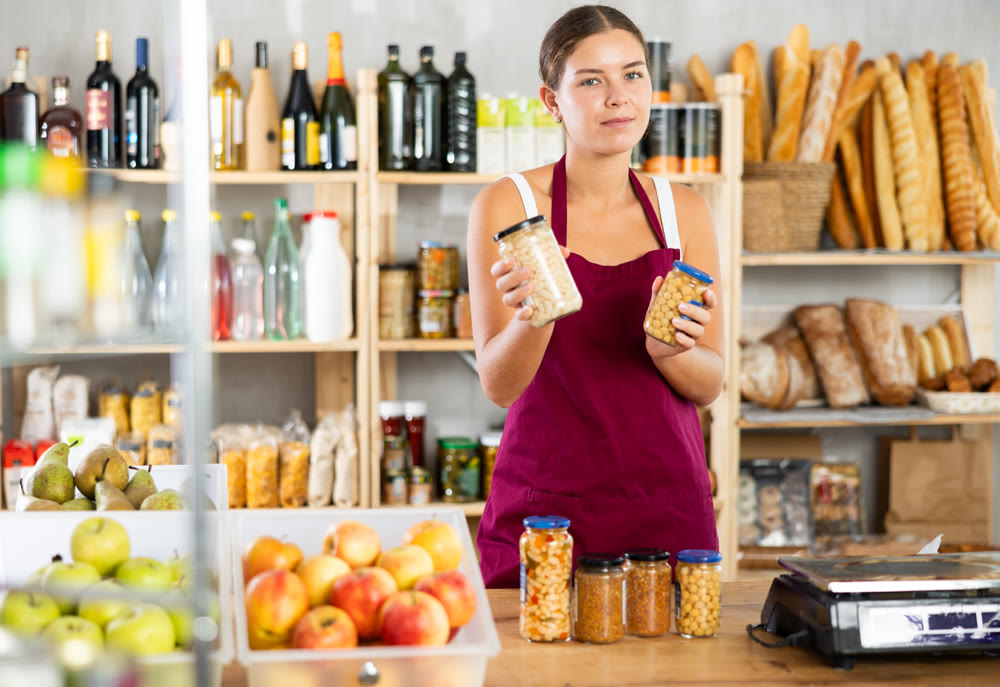 Portrait of friendly girl seller behind a store counter offering to buy canned beans and legumes