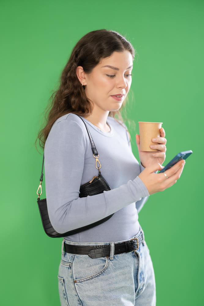Pretty young woman scrolling through her smartphone screen against and drinking coffee green screen background. Chroma key compositing