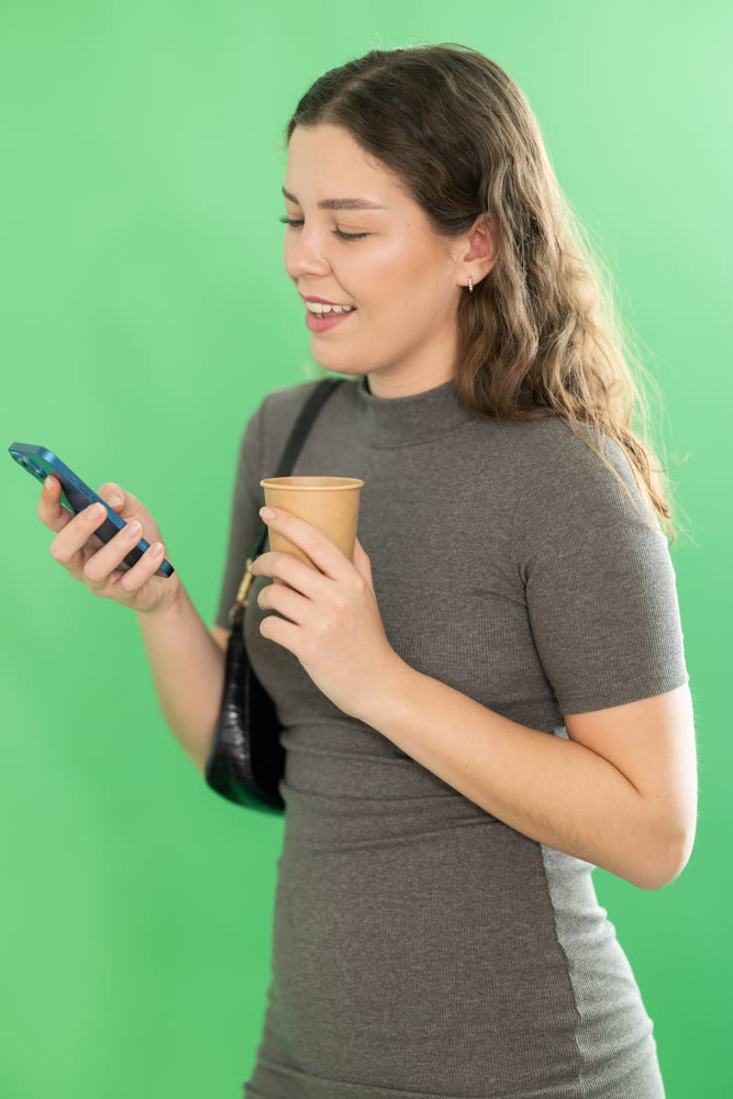 Young woman scrolling smartphone and drinking coffee in front of green screen