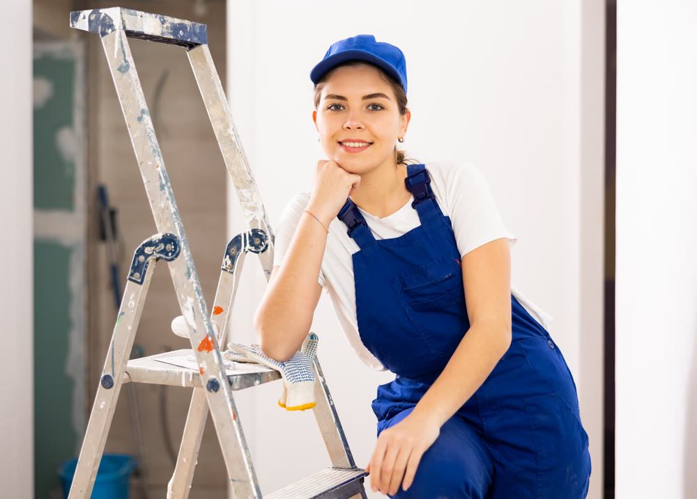 Portrait of positive builder woman in blue overalls next to stepladder