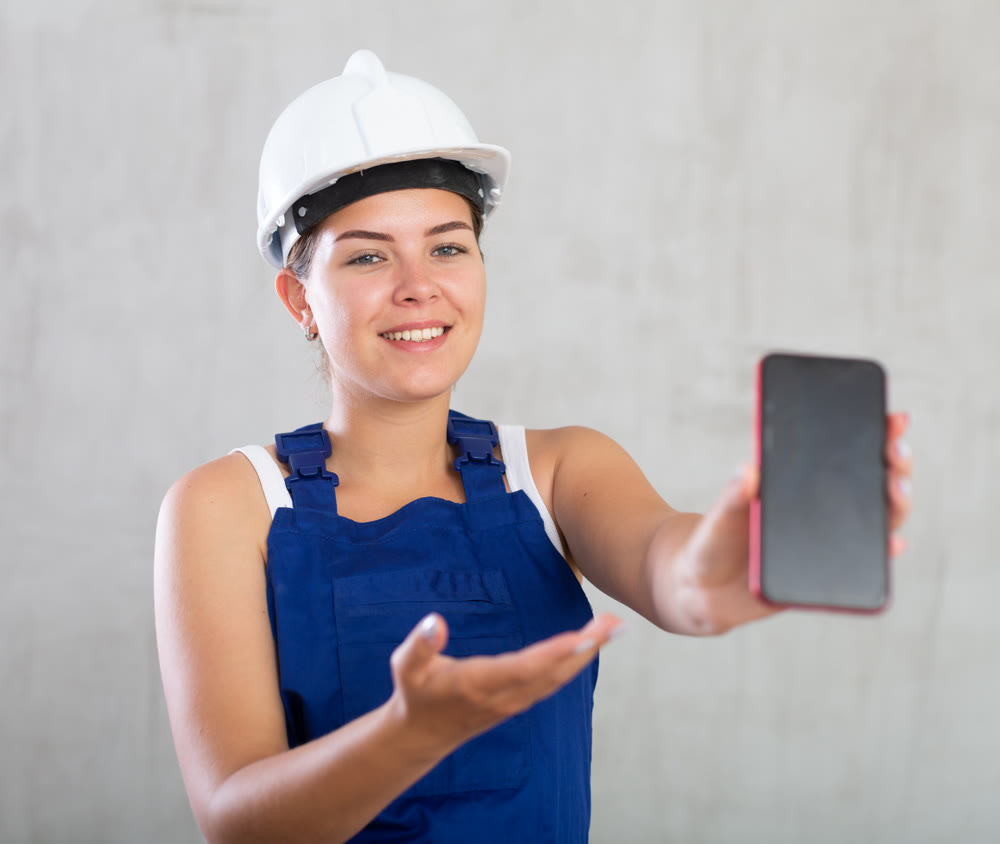 Young female engineer, architect in construction overalls and safety helmet showing mobile phone screen