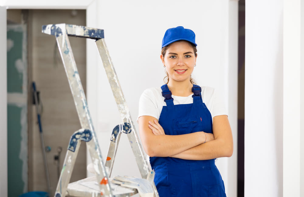 Portrait of positive builder woman in blue overalls next to stepladder