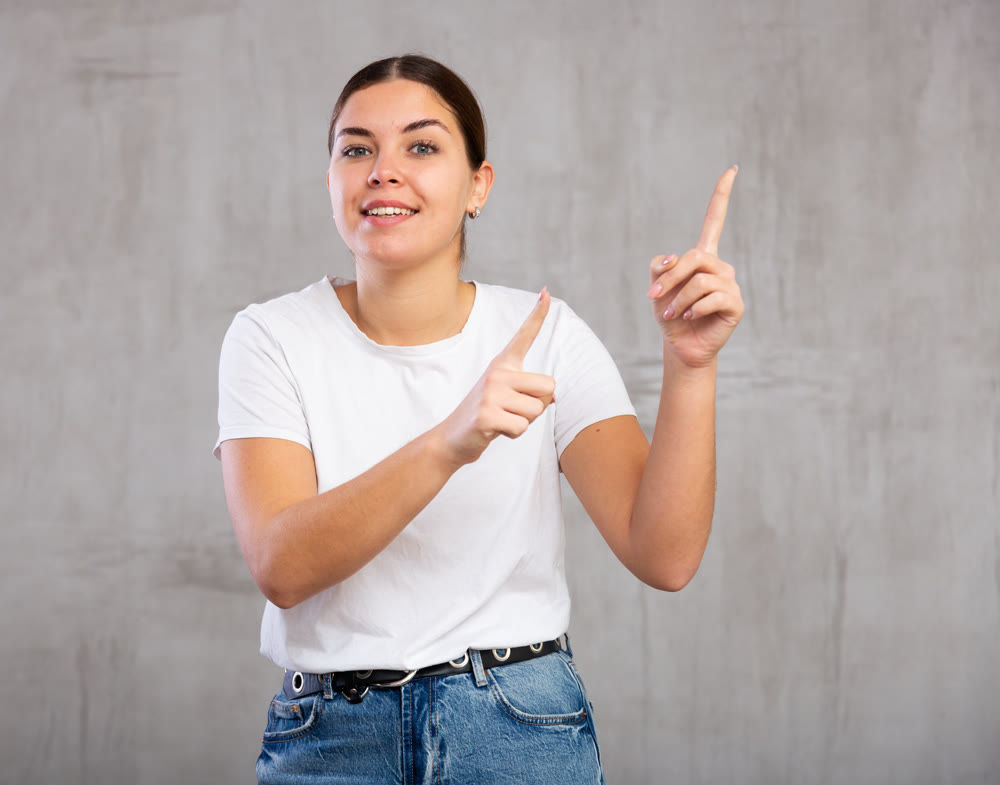Cheery young woman pointing fingers away against gray background