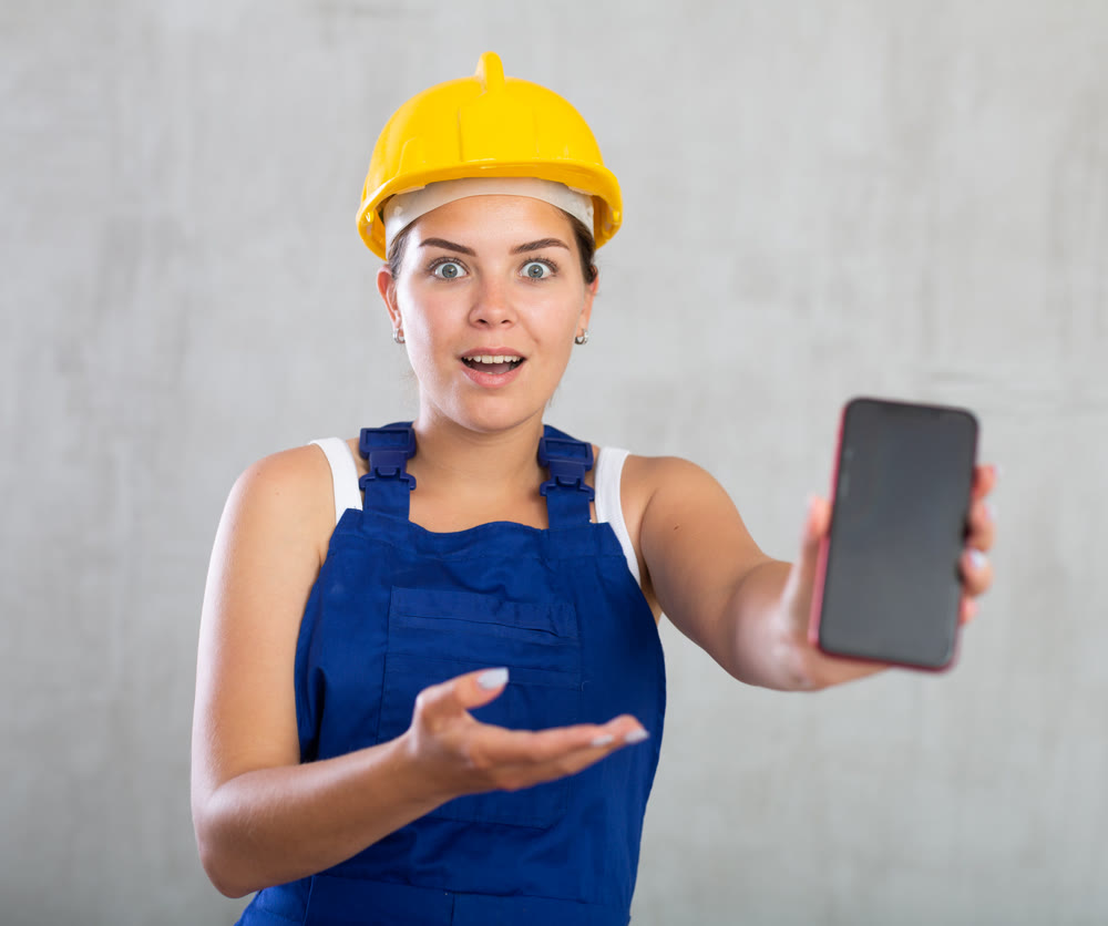 Smiling female worker in work clothes and hardhat demonstrating screen of her phone