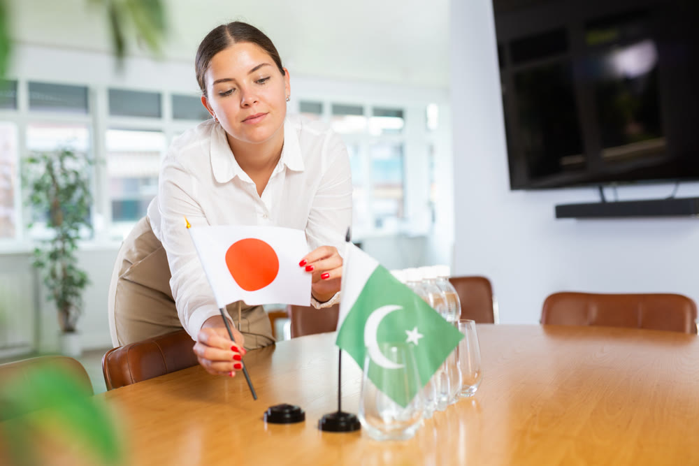 Positive young woman putting little flag of Japan on table next to the flag of Pakistan