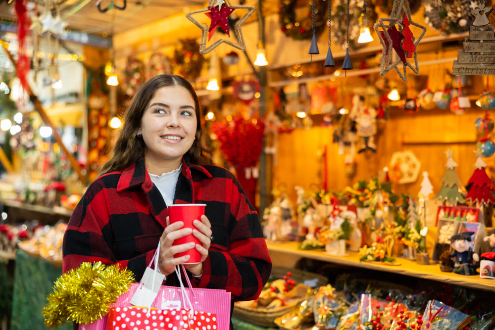 Girl walking in colorful Christmas street market, holding paper cup of warm mulled wine
