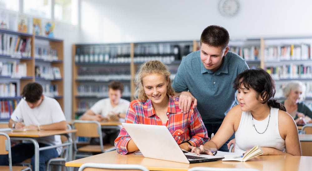 Smiling boy helping positive pupils research, working with laptop in library