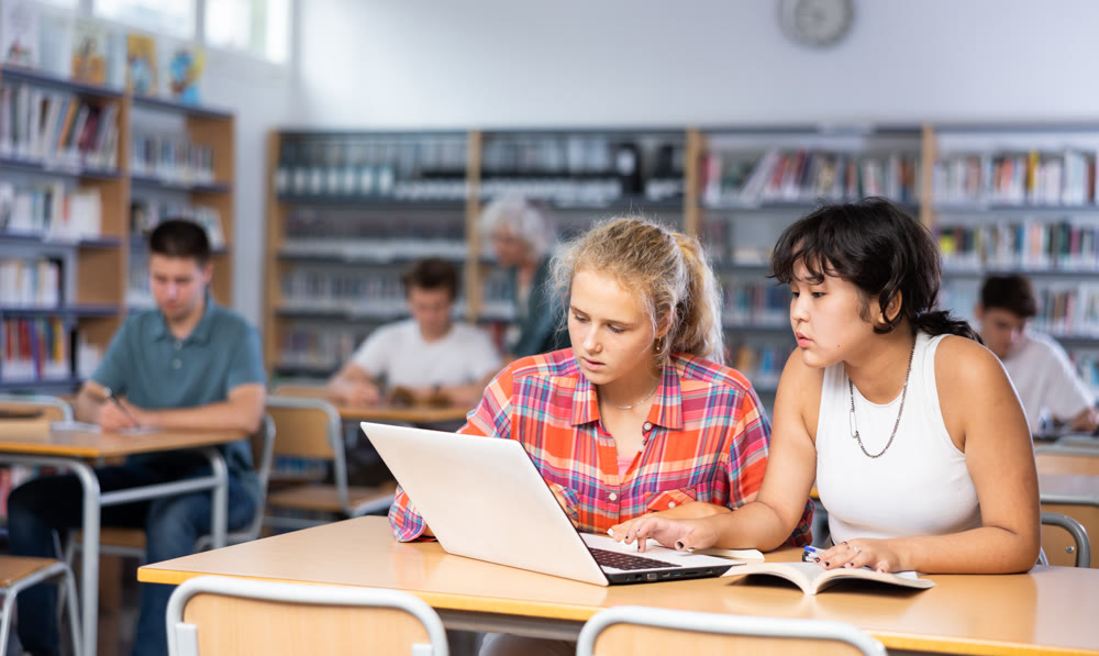 Two schoolgirls studying on a laptop and writing abstract in copybooks