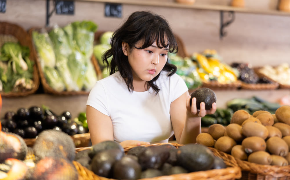 Interested young asian girl choosing ripe avocados in supermarket