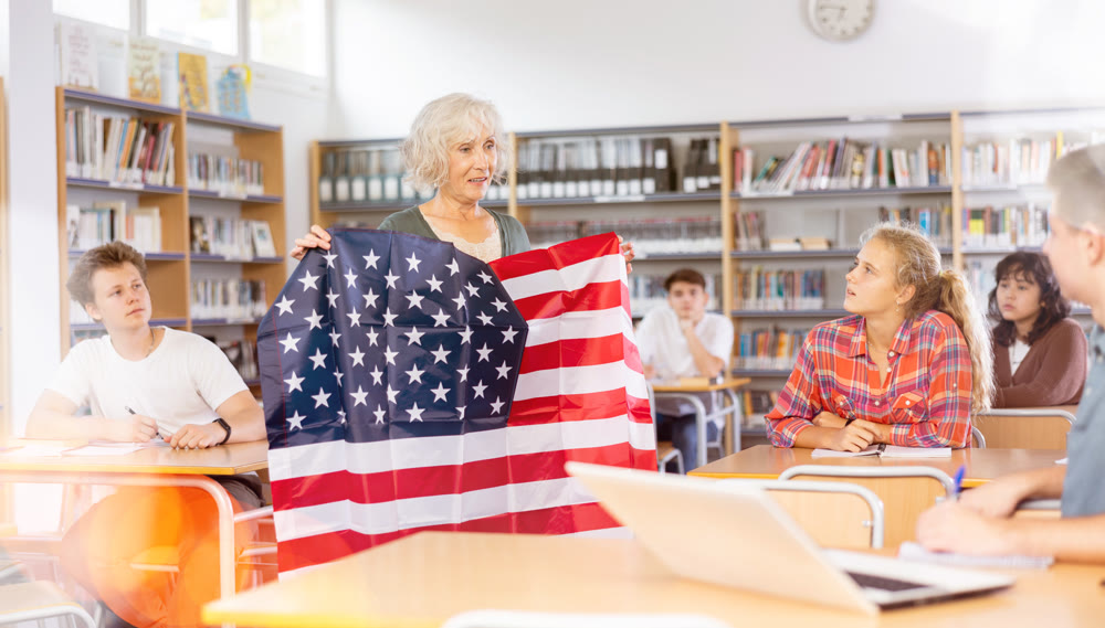 Mature female teacher shows the flag of United States of America to teenagers and talks about this country during lesson at library school