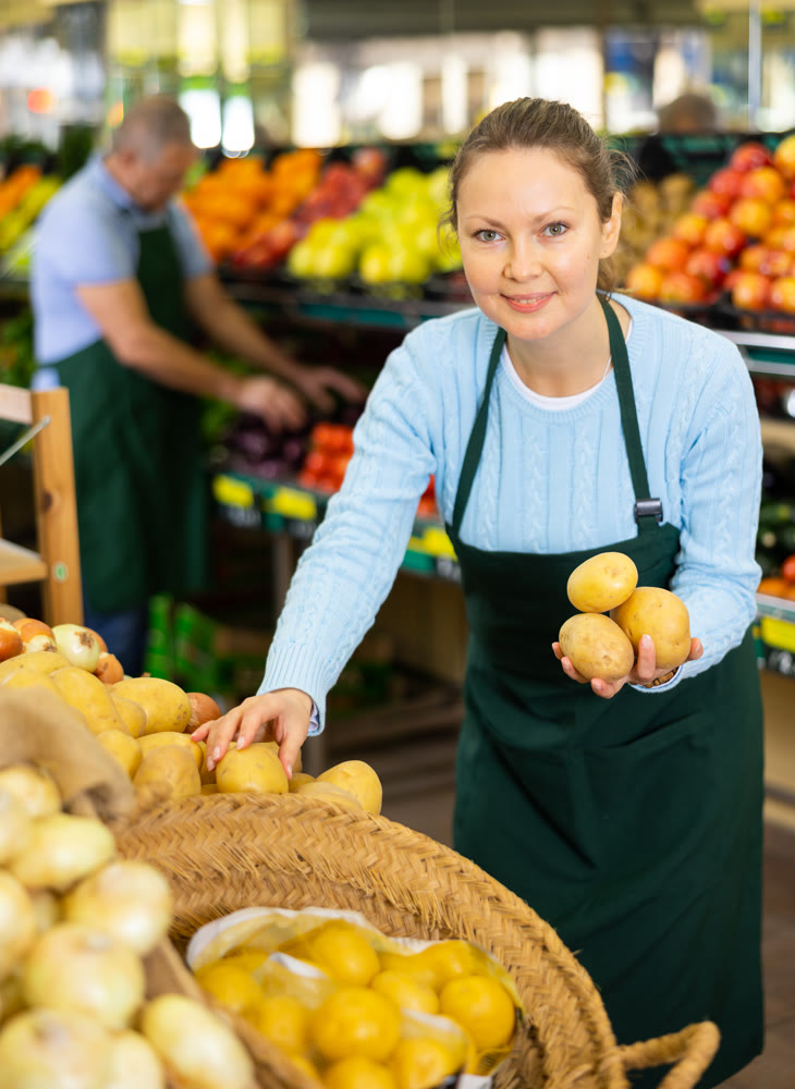 Friendly middle-aged woman market assistant arranging fresh potatoes in grocery supermarket