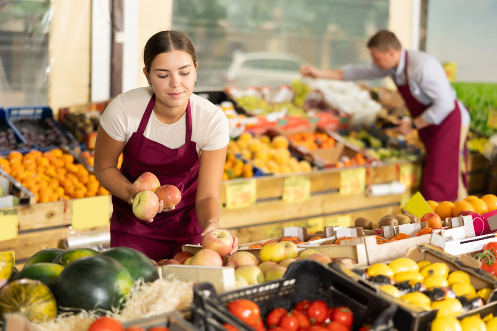 Young salesgirl laying out fresh apples for sale in greengrocery