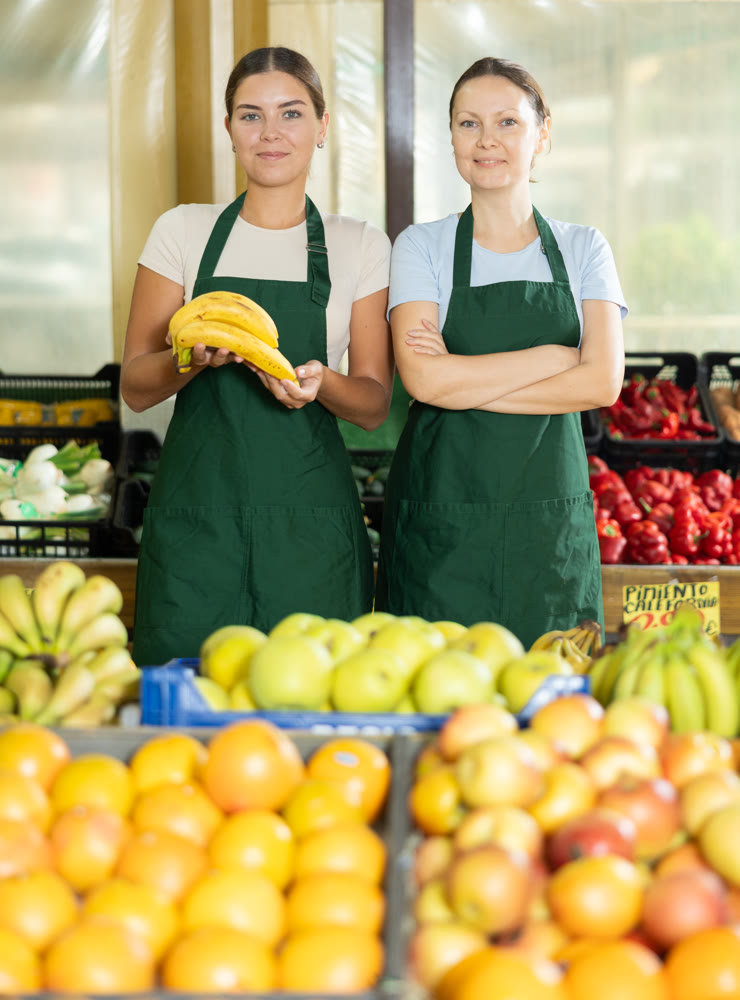 Happy two female sellers wearing apron and standing next to organic foods in supermarket