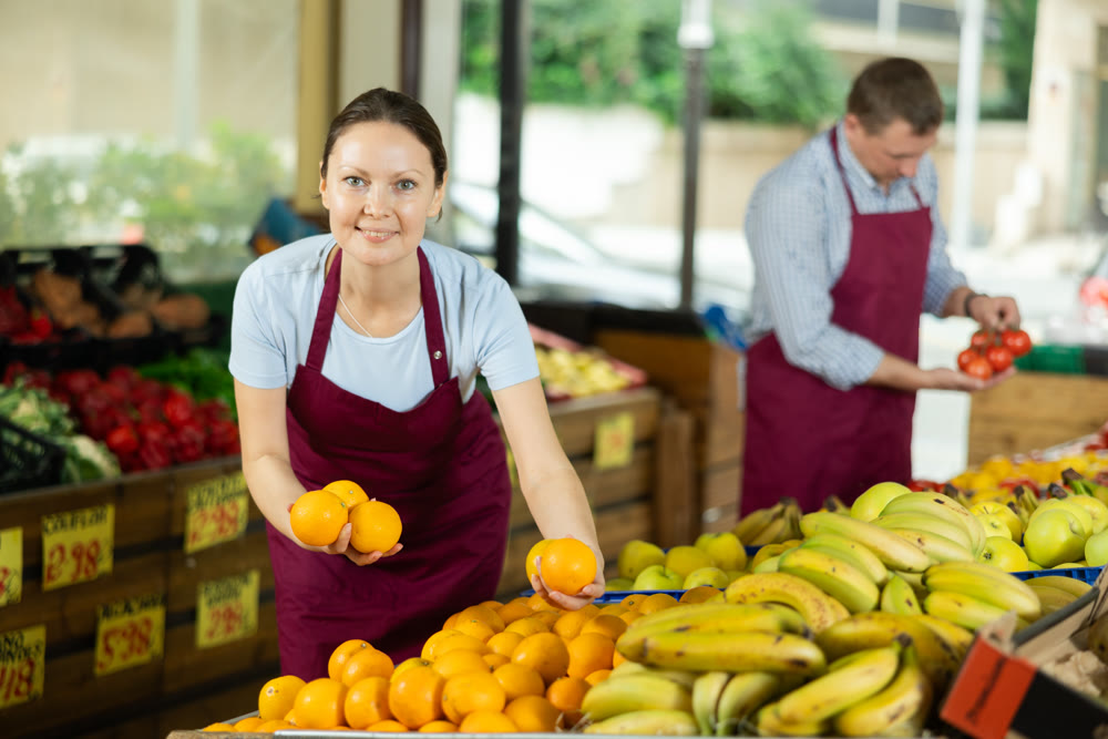 Woman selelr of vegetable department store is replenishing showcase with oranges