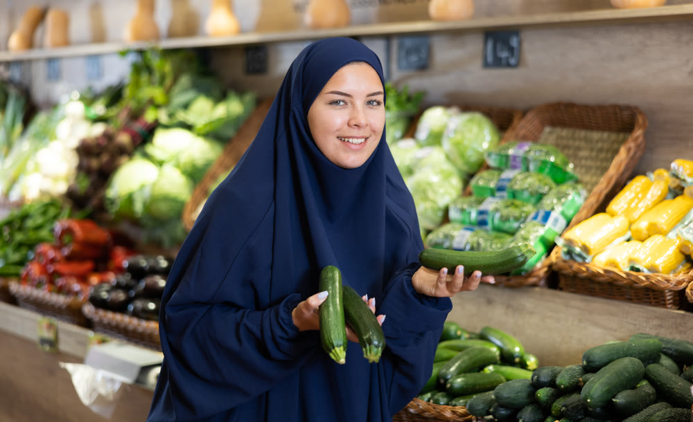Delighted young Muslim woman purchaser choosing cucumber in grocery store