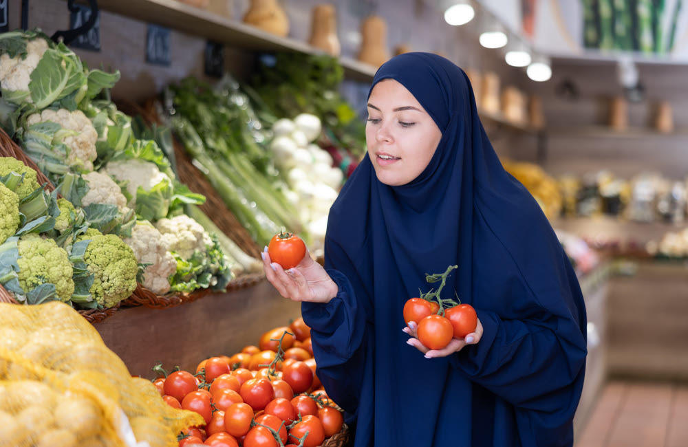 Young european girl in islamic dress choosing tomatoes in supermarket