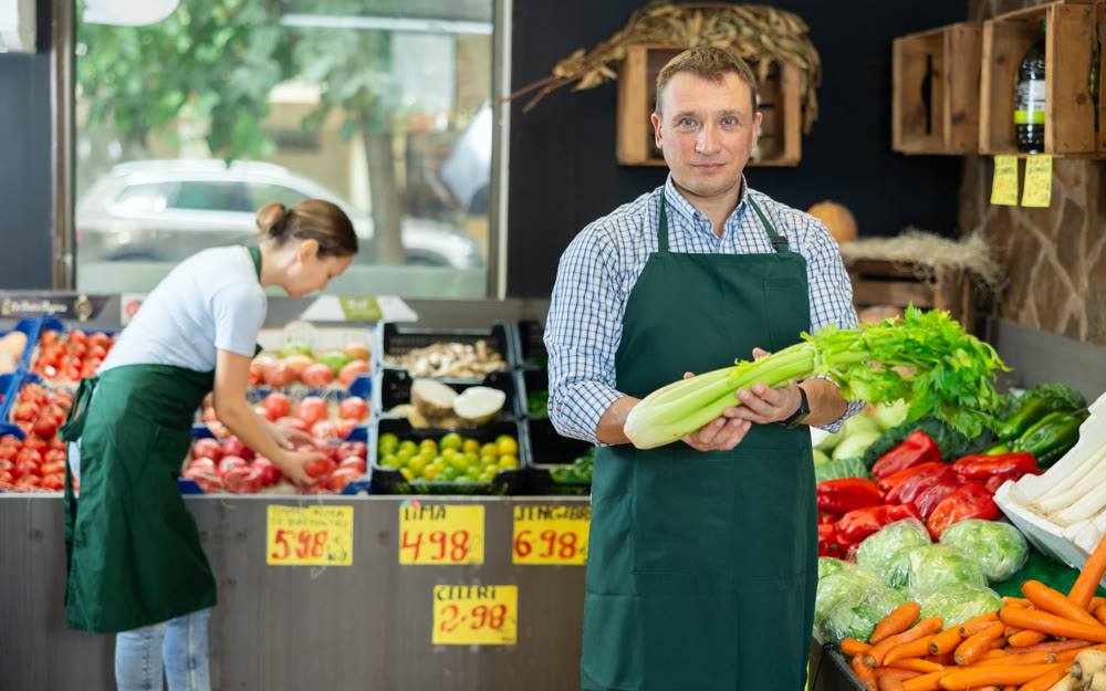 Man seller of fruit and vegetable store works near showcase, sorting greenery celery