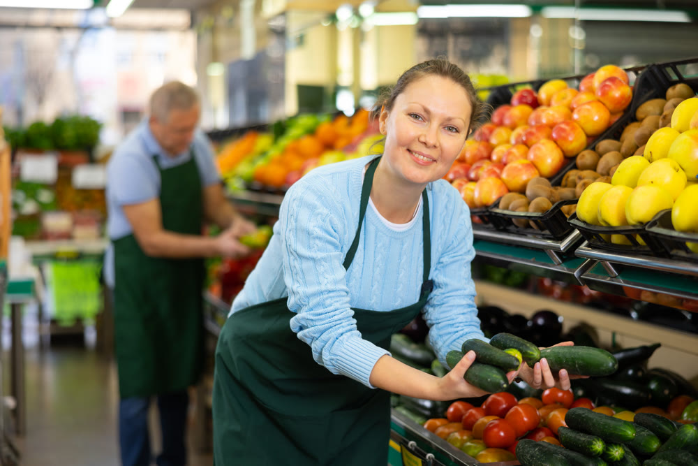 Middle-aged saleswoman putting cucumbers on food stall in greengrocery