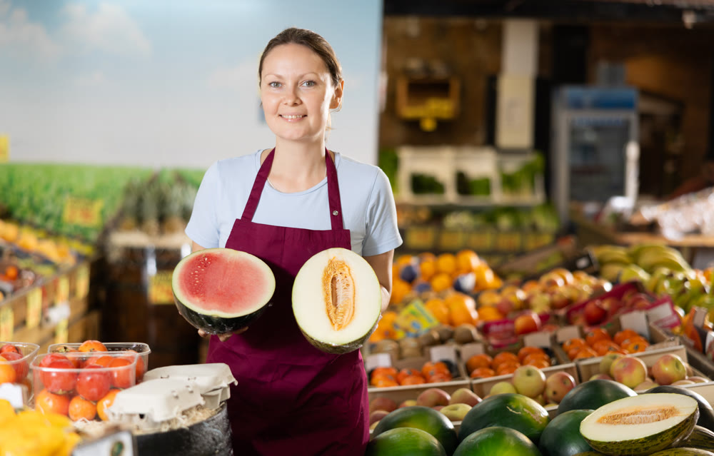 Woman grocery store seller offers a choice of buying ripe melon or watermelon.