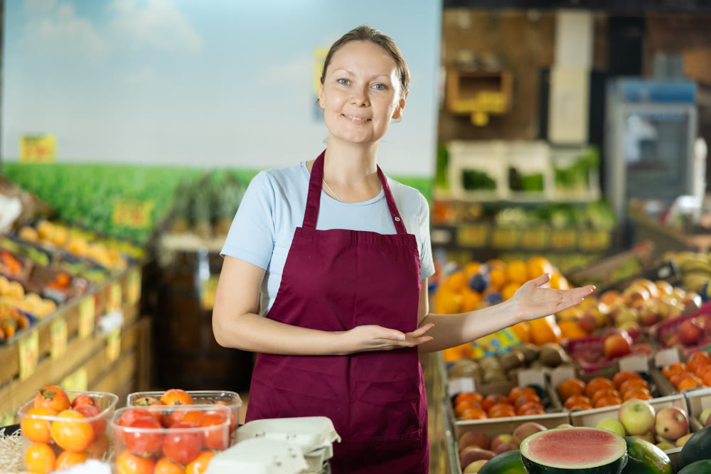 Portrait of saleswoman posing at grocery store. Invites you to go shopping