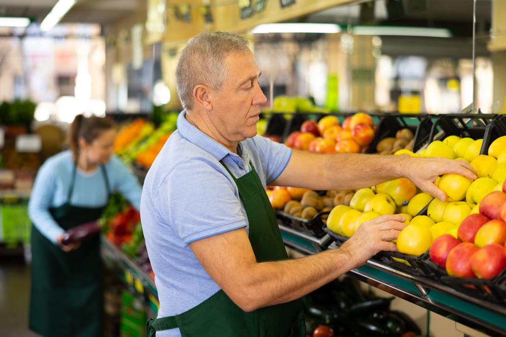 Focused elderly man in apron arranging Golden Delicious apples in fruit section