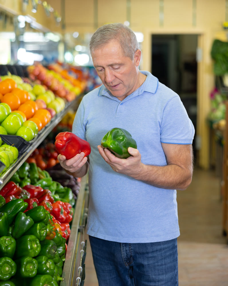 Focused elderly male pensioner in casual wear choosing local bell peppers during shopping in store