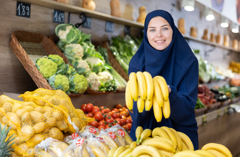 Girl in veil choosing fresh yellow bananas in fruit and vegetable section of supermarket