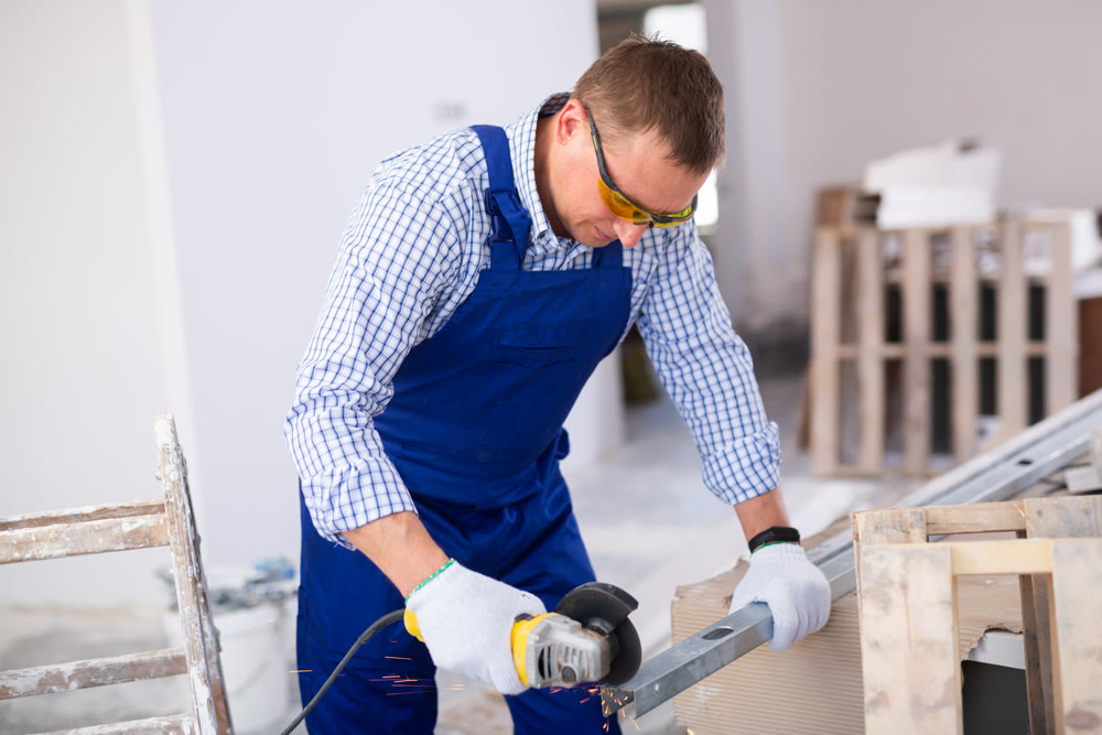 Professional builder in blue overalls cuts an aluminum profile with angle grinder