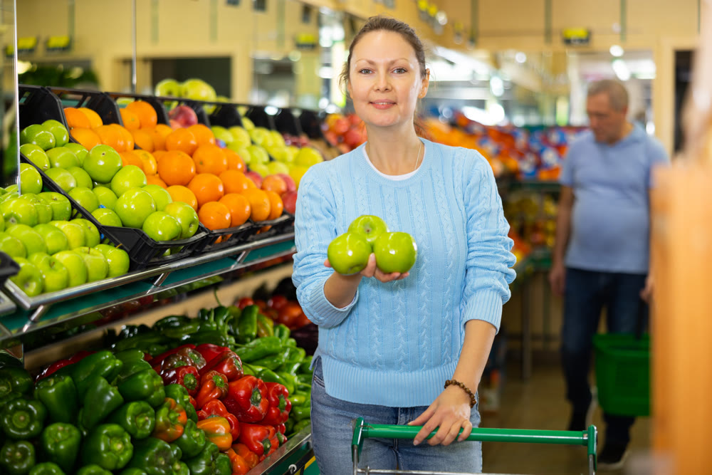Happy middle-aged female in casual clothes shopping and choosing green apples in supermarket