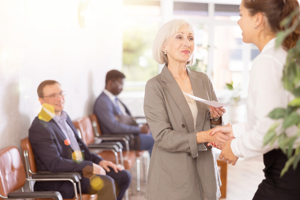 Smiling young girl makes deal, shaking hands with elderly woman colleague while standing in office.