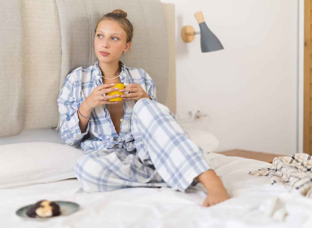 Young girl in pajamas drinking hot coffee and eating cookies while sitting on bed in bedroom - breakfast in bed