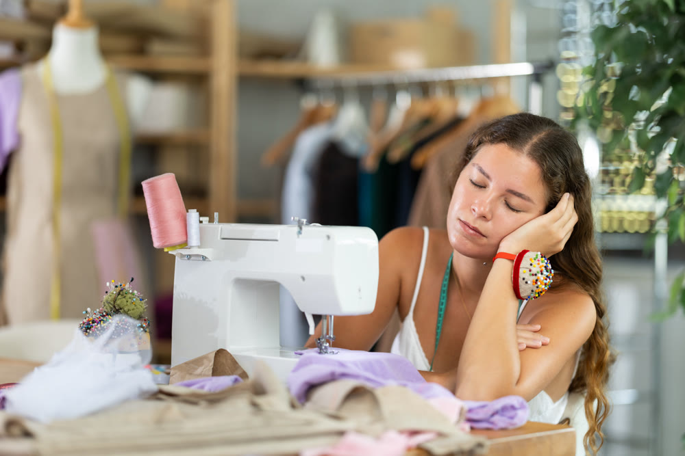 Exhausted young female tailor falling asleep at desk in workshop