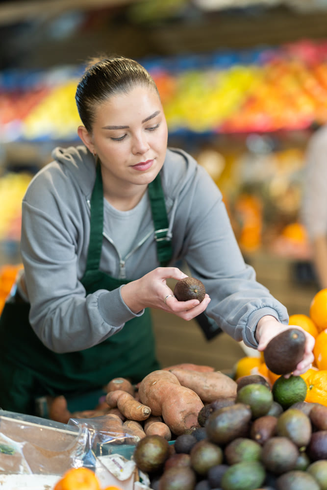 Young woman seller puts avocados in vegetable shop