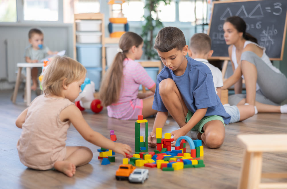 Little two boys sitting on floor together and playing educational games and toys at classes for kids, other children in background