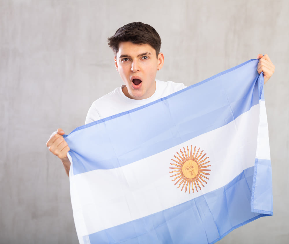 Happy young man holding flag of Argentina against unicoloured background