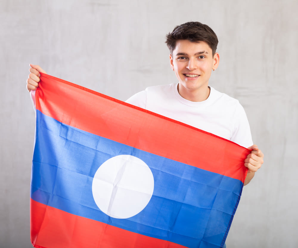Happy young man holding flag of Laos against unicoloured background